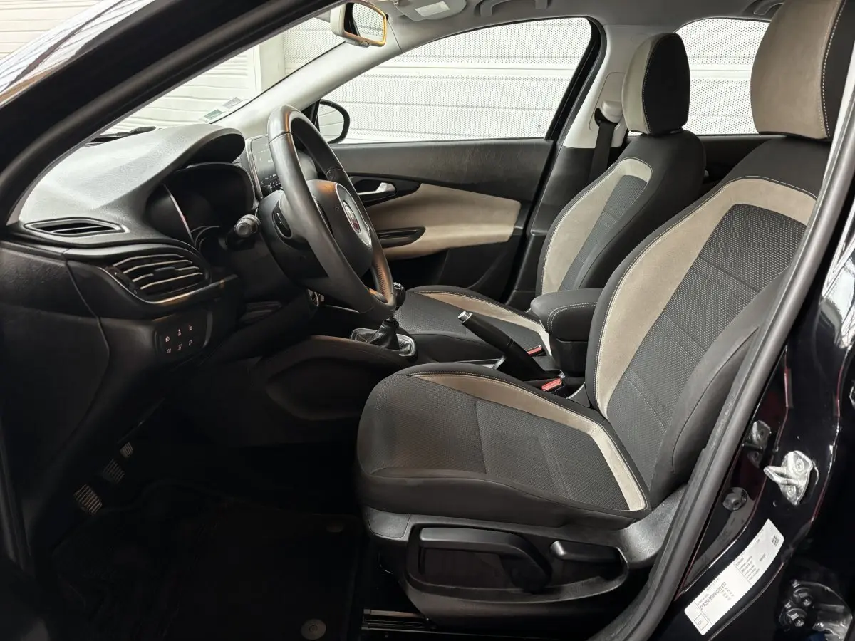Vue intérieure côté conducteur de la Fiat Tipo noire, montrant les sièges bi-matière et le volant multifonctions cuir alu.