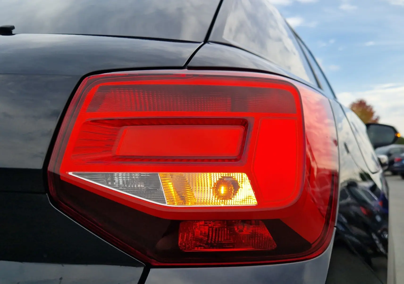 Gros plan sur le feu arrière droit rouge et orange d’un Audi Q2 noir mito metal 2023, avec reflet du ciel et voitures alentour.