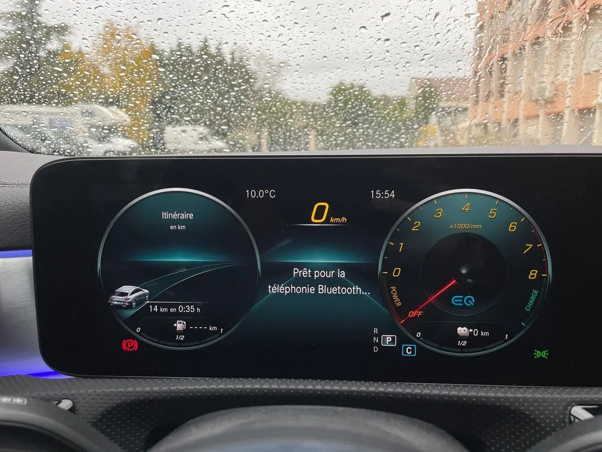 Tableau de bord numérique de Mercedes CLA Shooting Brake blanc, affichage du compteur et navigation sous la pluie.