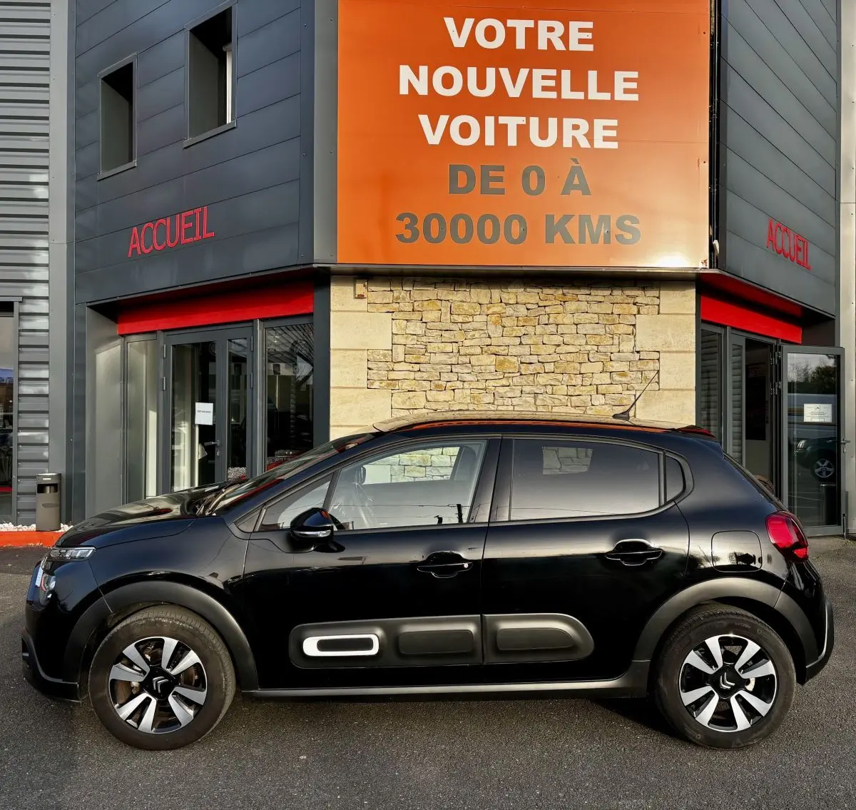 Profil latéral droit d'une Citroën C3 noire 2024 avec protections latérales blanches et jantes bicolores, devant un bâtiment commercial.