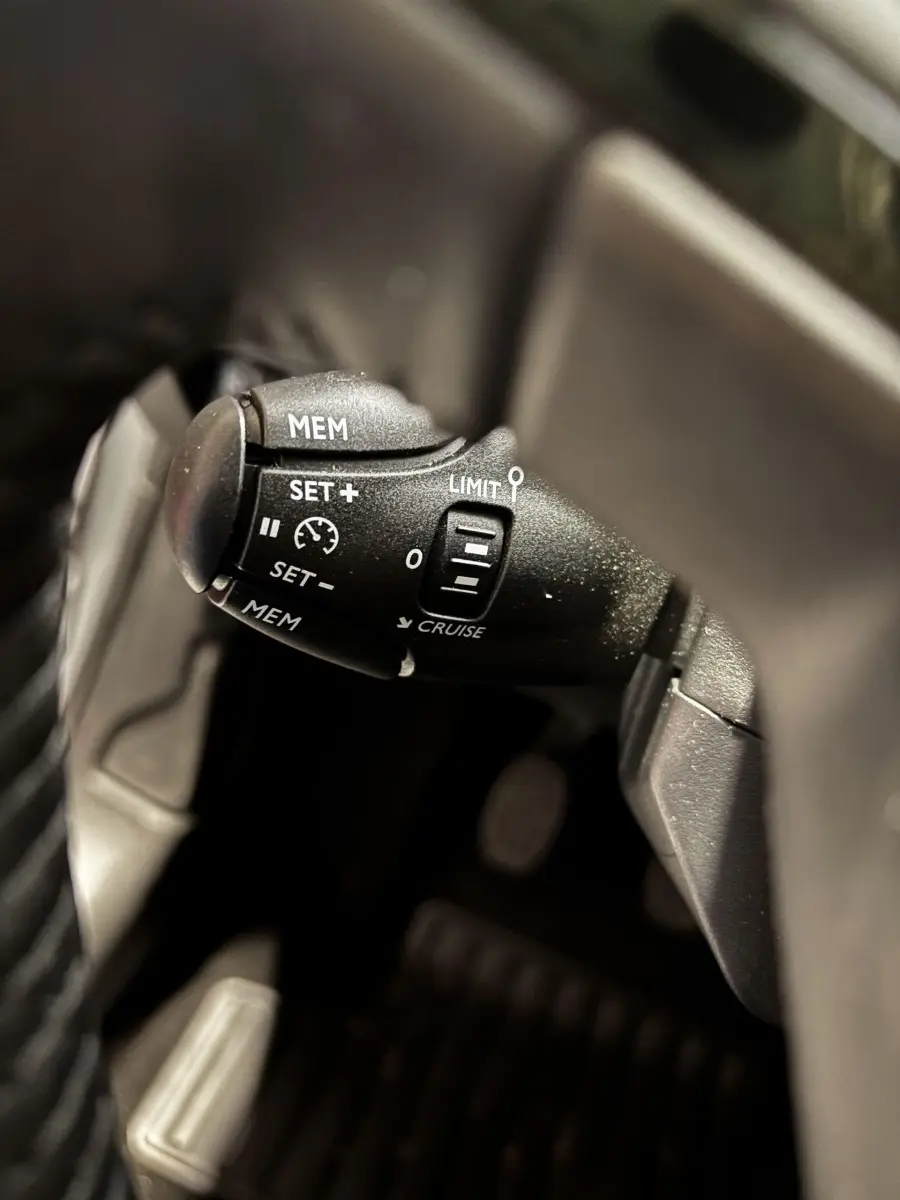 Gros plan sur la commande de régulateur de vitesse noire à gauche du volant d'une Peugeot 208 gris clair 2024.