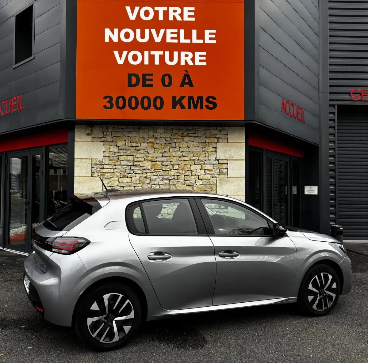 Vue latérale droite d'une Peugeot 208 PureTech 100 S&S Active gris clair stationnée devant un bâtiment avec panneau orange.