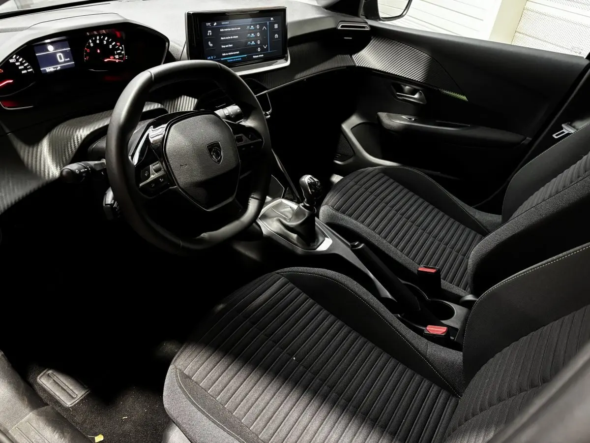 Vue intérieure côté conducteur de la Peugeot 208 PureTech 100 S&S Active 2024 avec tableau de bord numérique et sièges noirs.