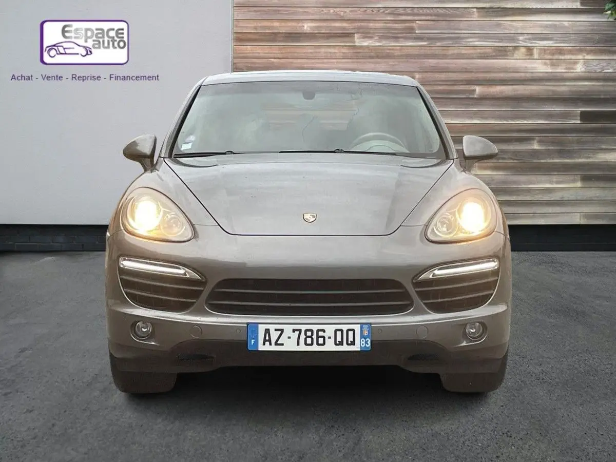 Vue frontale d'un Porsche Cayenne S Hybrid gris 2010 avec phares allumés et plaque d'immatriculation française visible.
