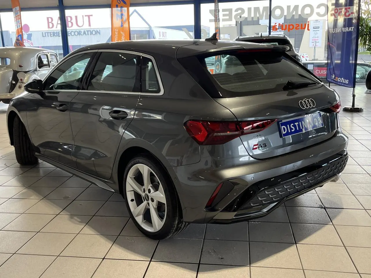 Audi A3 Sportback gris métallisé vue 3/4 arrière droit en intérieur avec jantes alliage et toit ouvrant panoramique.
