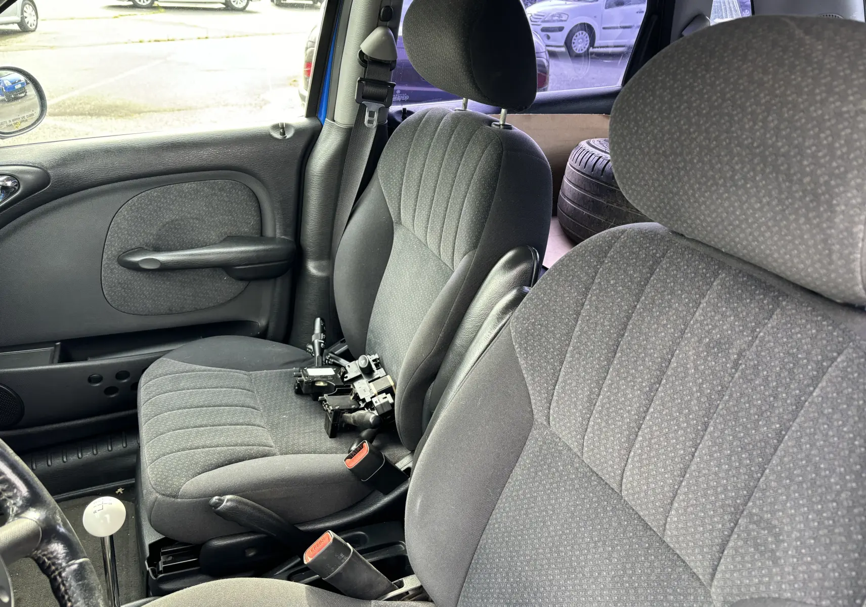 Vue intérieure côté gauche du Chrysler PT Cruiser bleu de 2004, montrant les sièges avant en tissu gris et une pièce mécanique posée.