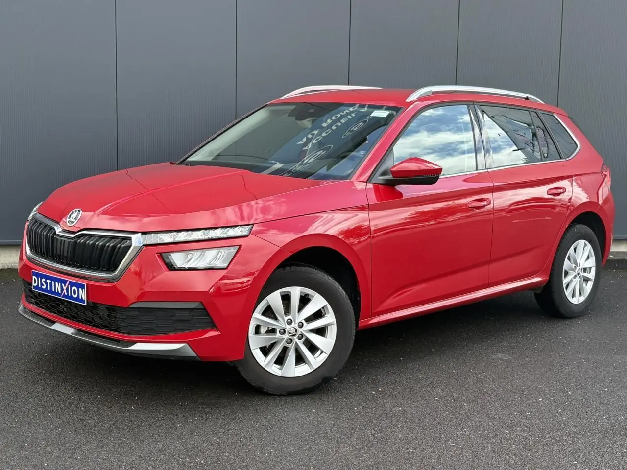 Vue 3/4 avant gauche d'un Skoda Kamiq TSI 110 DSG Ambition rouge Velvet avec jantes alliage 16 pouces et barres de toit alu.