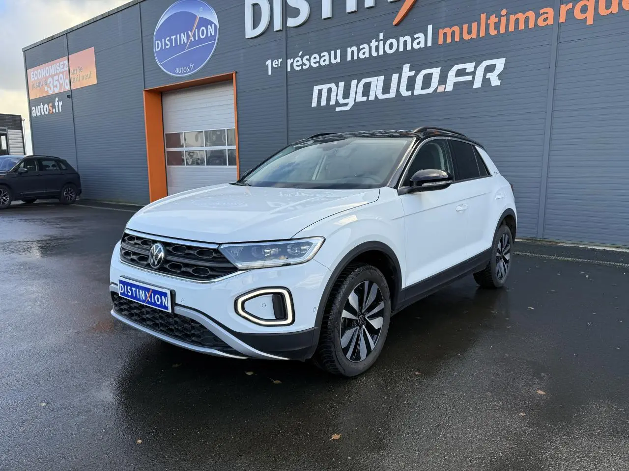 Volkswagen T-Roc blanc pur en 3/4 avant droit avec toit noir et jantes alliage 'Zürich' sur parking humide.