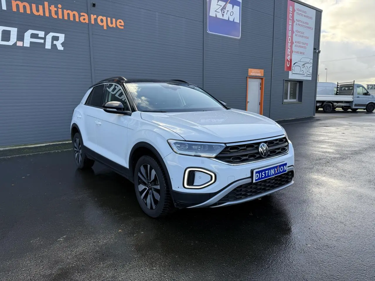 Volkswagen T-Roc blanc pur en 3/4 avant droit, avec jantes alliage et calandre noire devant un bâtiment gris.