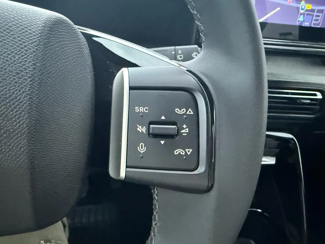 Gros plan sur les commandes gauche du volant noir de la Citroën C3 2025, avec tableau de bord et écran tactile en arrière-plan.