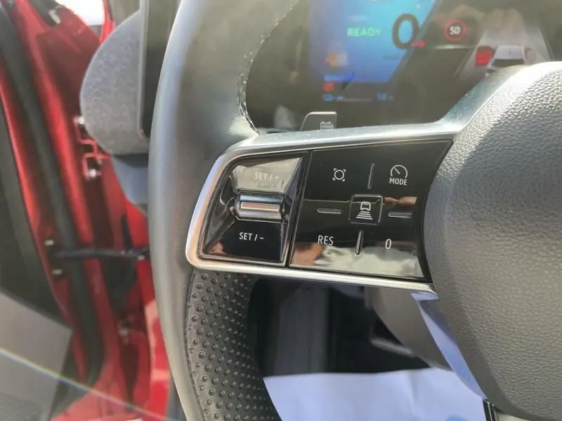 Gros plan sur les commandes gauche du volant du Renault Scénic E-Tech rouge flamme 2024, avec tableau de bord numérique en arrière-plan.