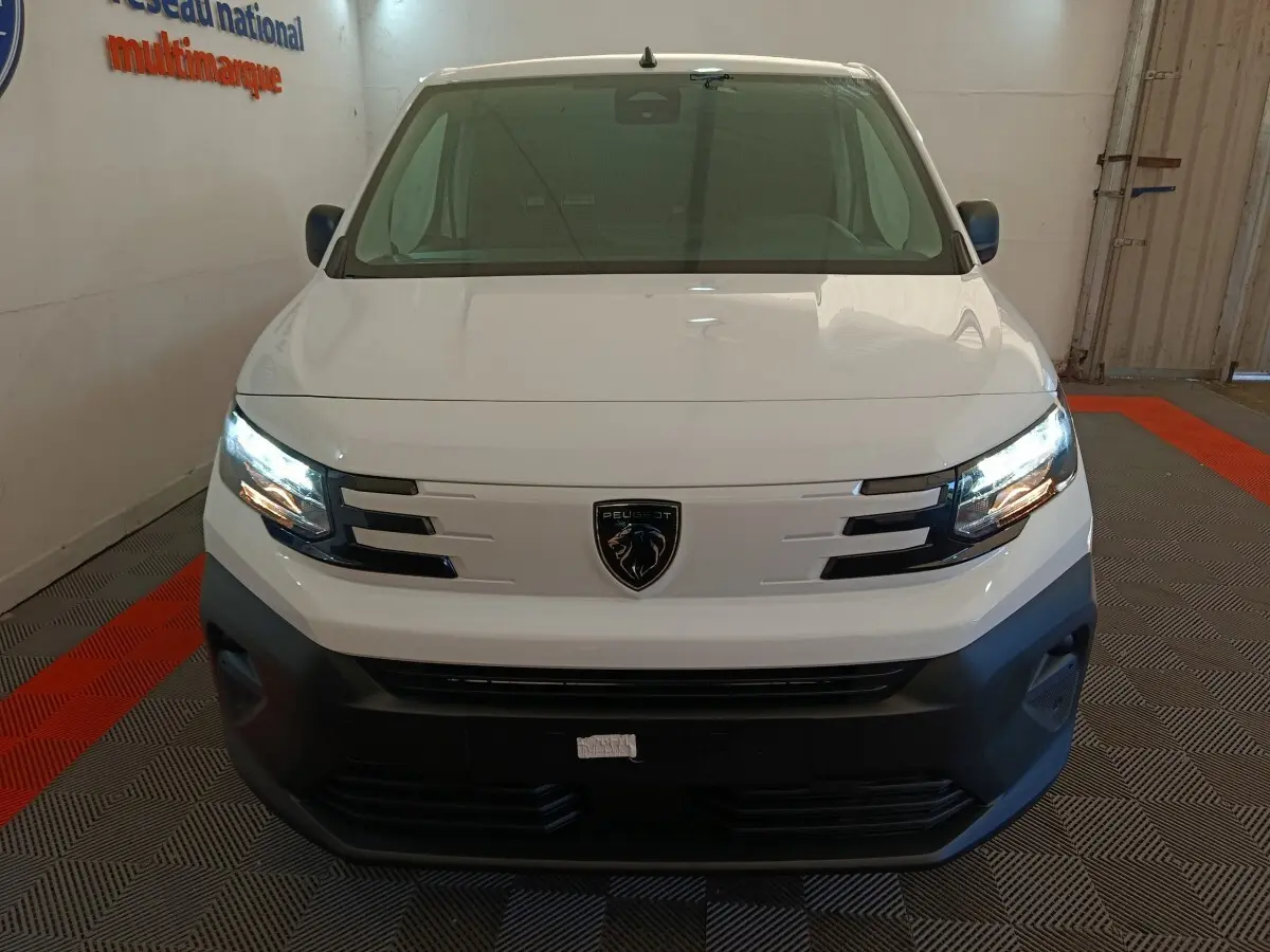 Vue de face du Peugeot Partner blanc 2025 avec phares allumés et calandre noire distinctive dans un garage.