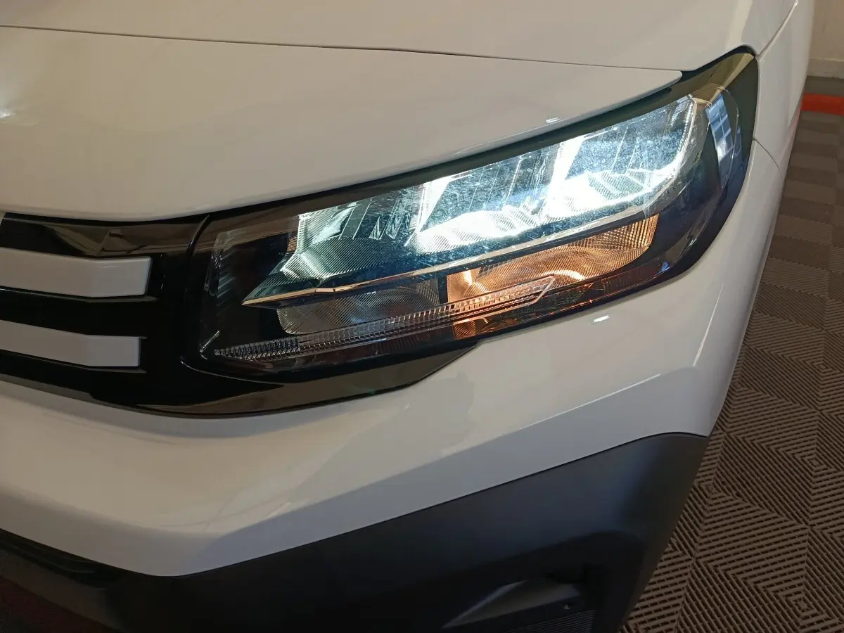 Gros plan sur le phare avant droit du Peugeot Partner blanc 2025, avec calandre noire et détails lumineux distinctifs.