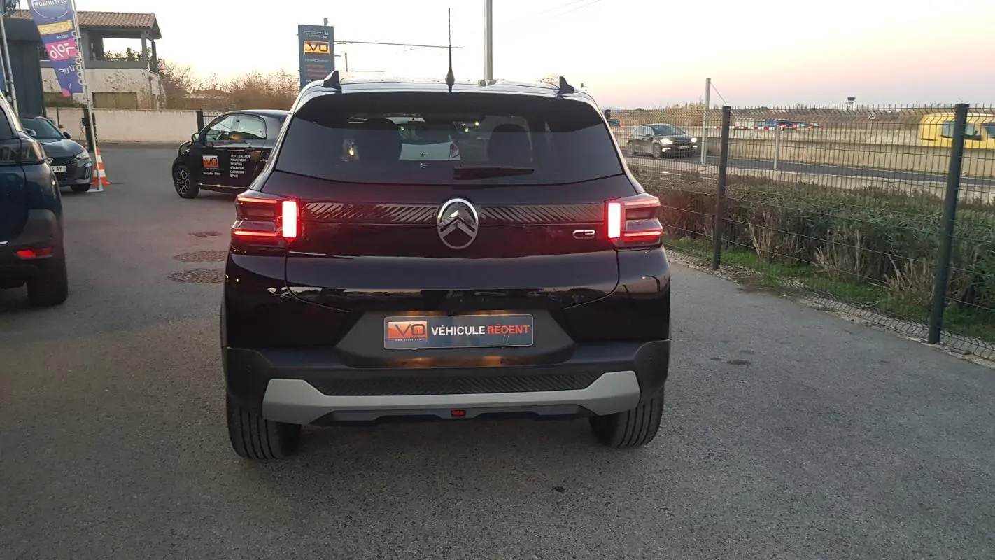 Vue arrière d'une Citroën C3 noire 2025 avec feux allumés et barres de toit visibles sur un parking extérieur.
