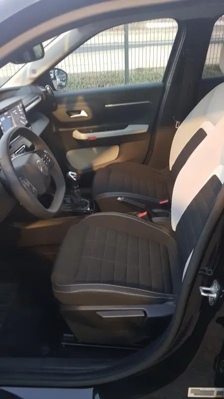 Vue intérieure côté conducteur de la Citroën C3 2025 noire, sièges bicolores noirs et blancs avec volant et console visibles.