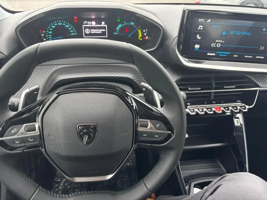Vue intérieure centrée sur le volant noir d'une Peugeot 208 Hybrid 2025 avec tableau de bord numérique et écran tactile.