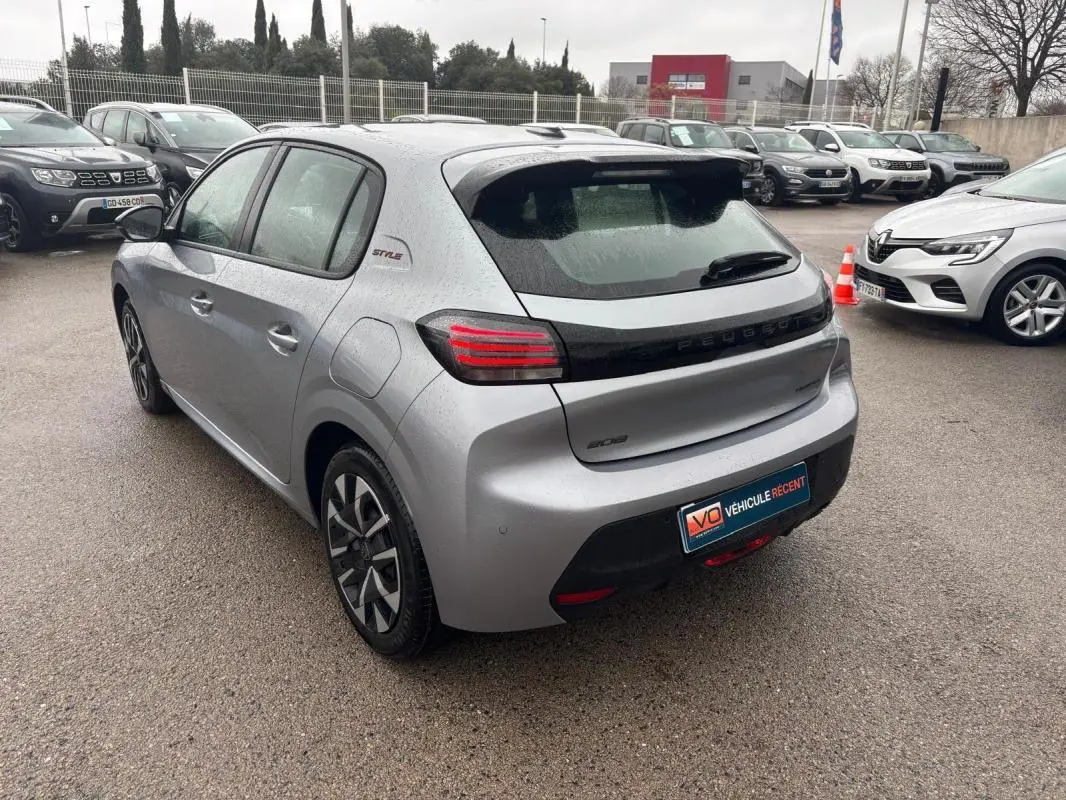 Vue 3/4 arrière gauche d'une Peugeot 208 Hybrid gris métallisé avec jantes 16 pouces et badge Style sous la pluie.