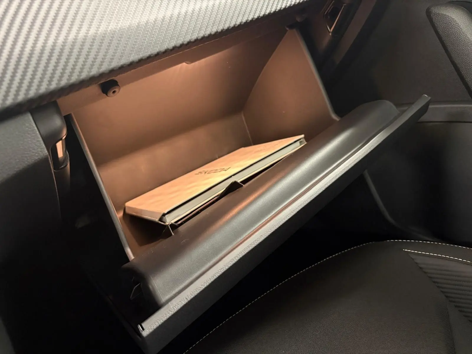 Gros plan sur la boîte à gants ouverte du Skoda Kamiq 2024 Monte Carlo, avec manuel à l'intérieur et sellerie noire visible.