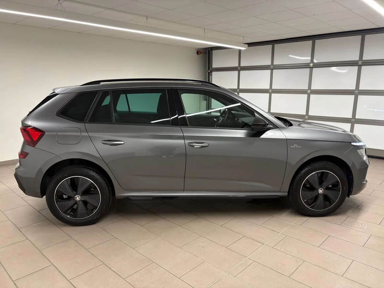 Vue de profil côté gauche d’un Skoda Kamiq 1.5 TSI Monte Carlo gris graphite avec jantes noires distinctives, en intérieur.