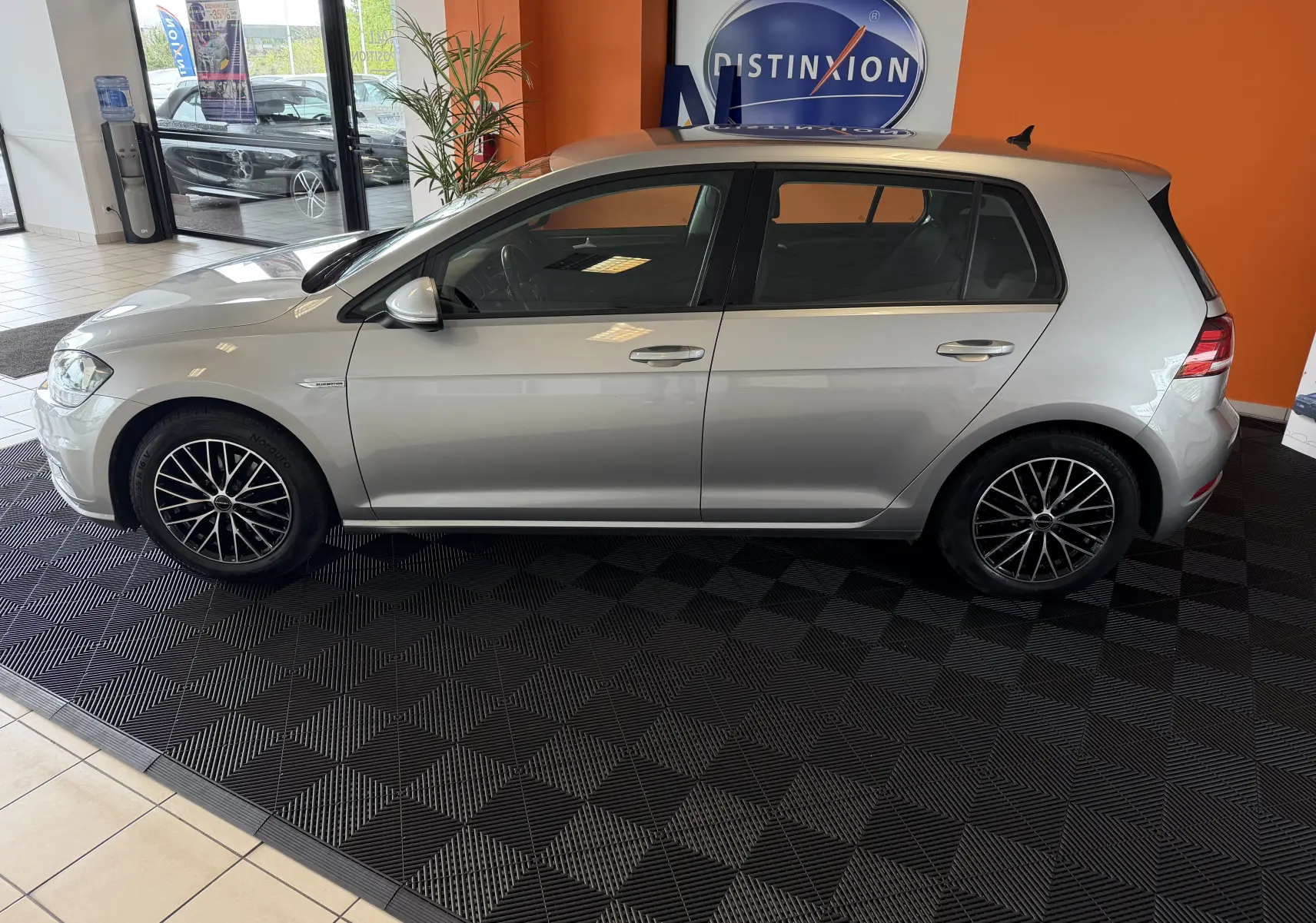 Profil côté gauche d'une Volkswagen Golf TSI 130 Confortline gris métallisé, avec jantes alliage noires, en intérieur showroom.