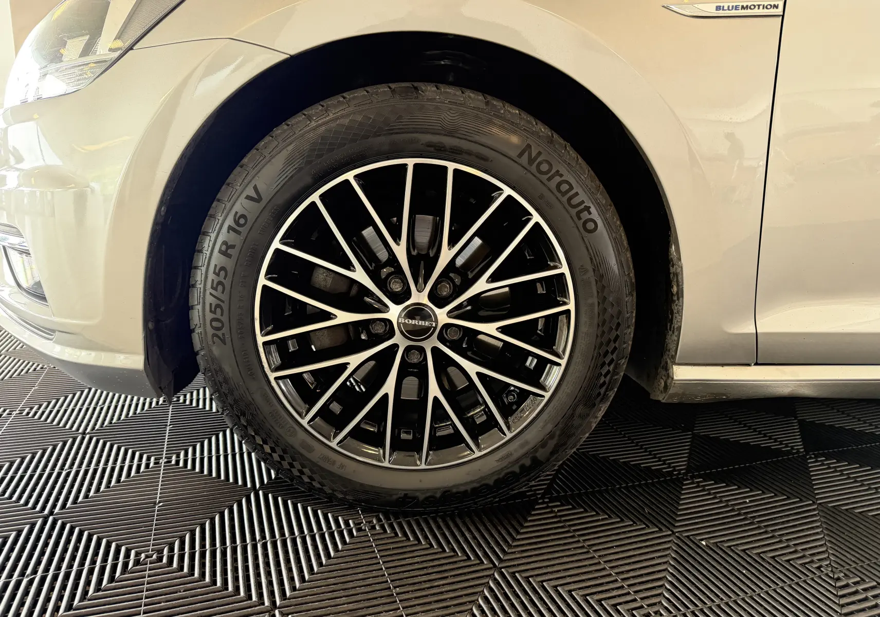 Gros plan sur la roue avant gauche d'une Volkswagen Golf gris clair avec jante alliage noir et argent et badge Bluemotion sur l'aile.