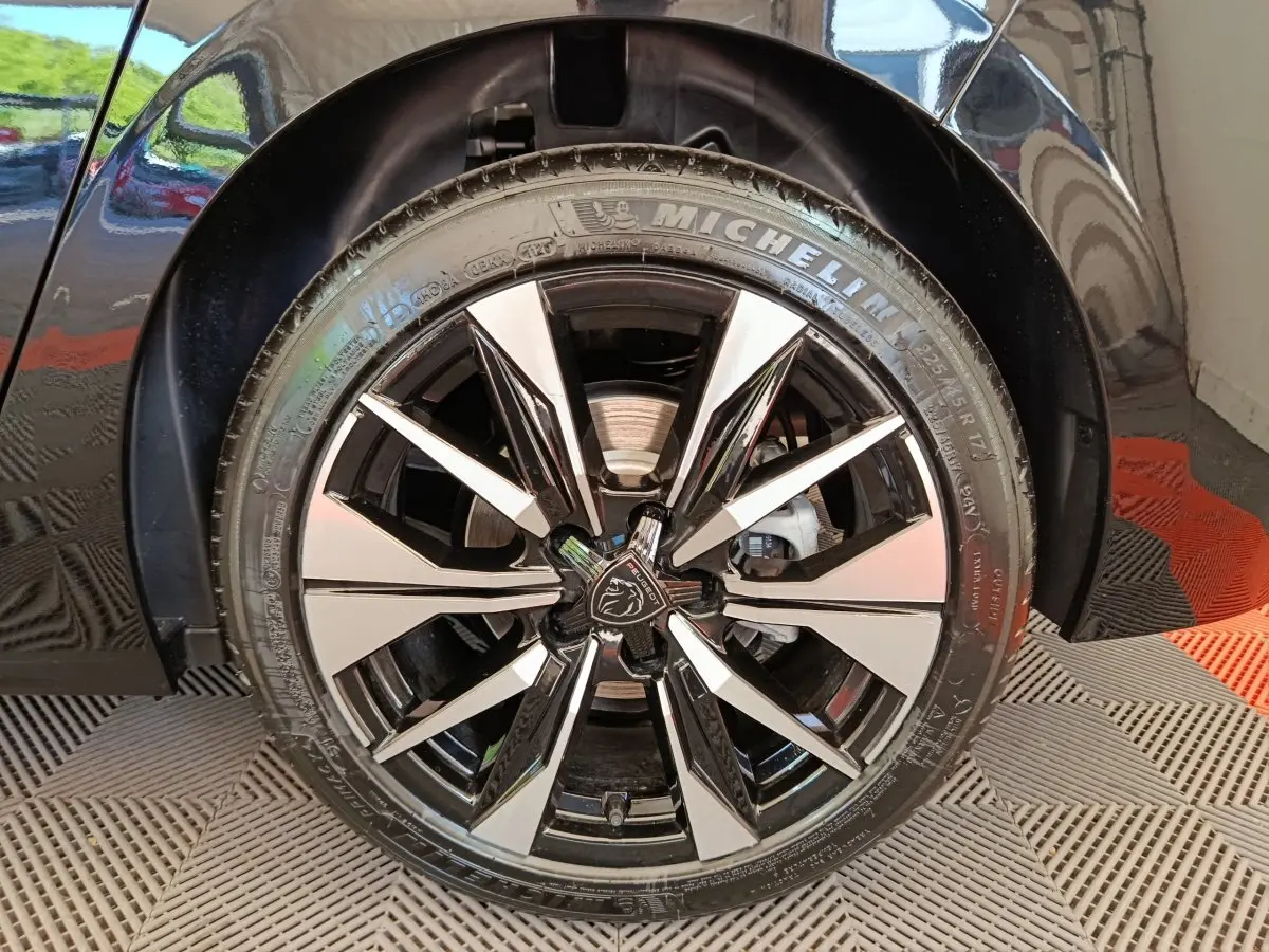 Gros plan sur la roue avant droite noire d'une Peugeot 308 2026 avec jante alliage bicolore et logo lion central.