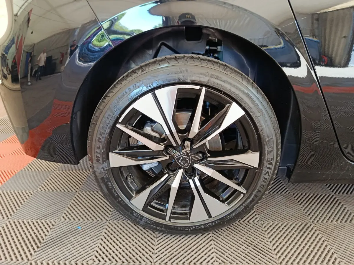 Gros plan sur la roue avant droite noire d’une Peugeot 308 2026 avec jante alliage bicolore et logo central lion.