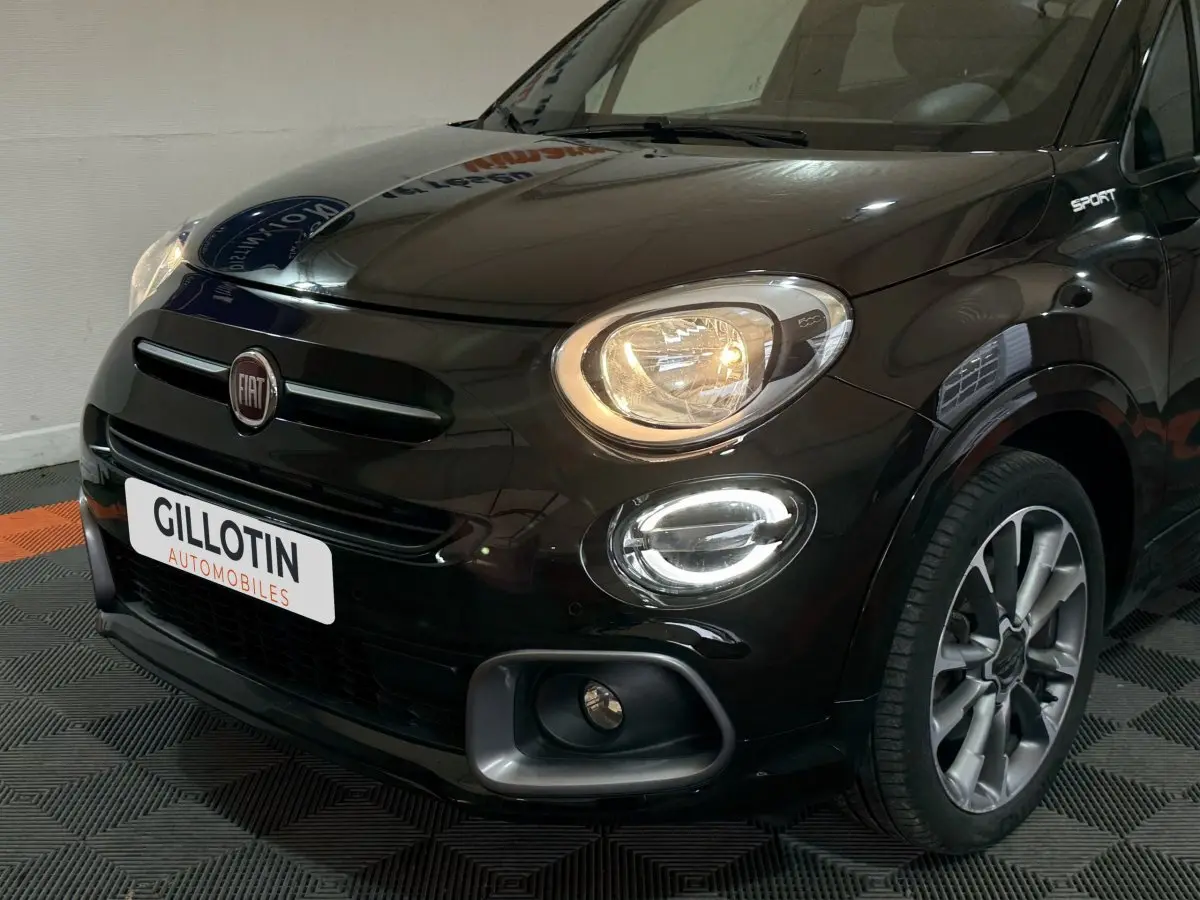 Vue 3/4 avant droit d'une FIAT 500X noire avec phares allumés et logo Sport visible sur l'aile avant.