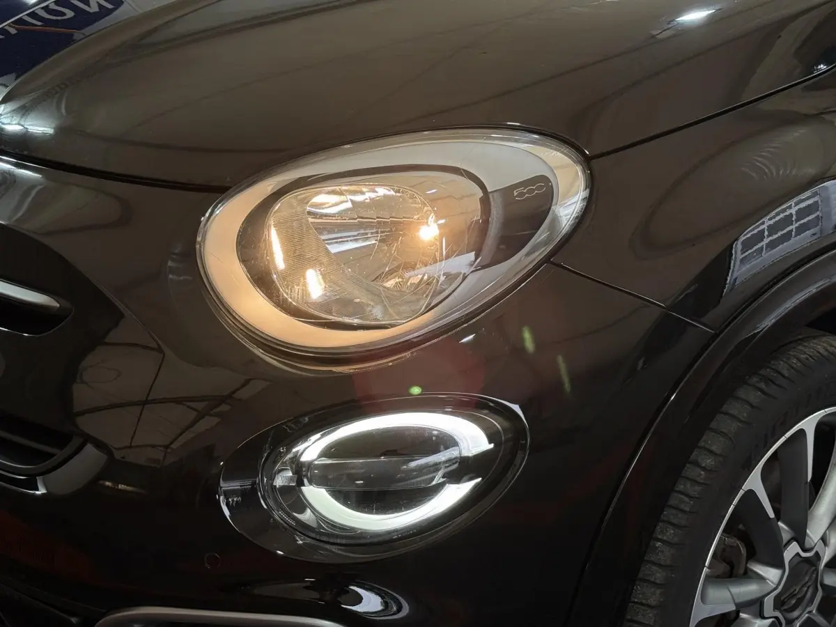 Gros plan sur le phare avant gauche allumé et le feu antibrouillard LED d'une FIAT 500X noire, avec jante visible.