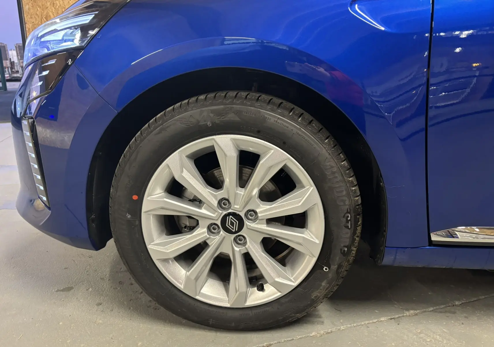 Gros plan sur la roue avant gauche de la Renault Clio bleu iron 2024 avec jante argentée et partie avant du véhicule visible.