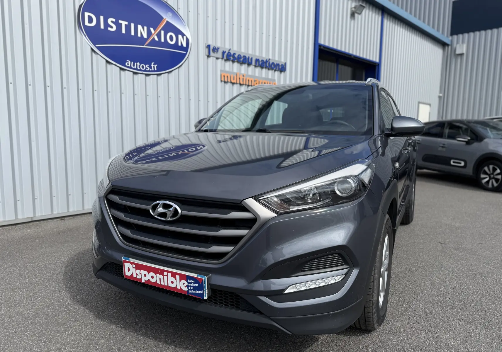 Hyundai Tucson gris en 3/4 avant droit, avec calandre marquée et feux LED, stationné devant un bâtiment commercial.