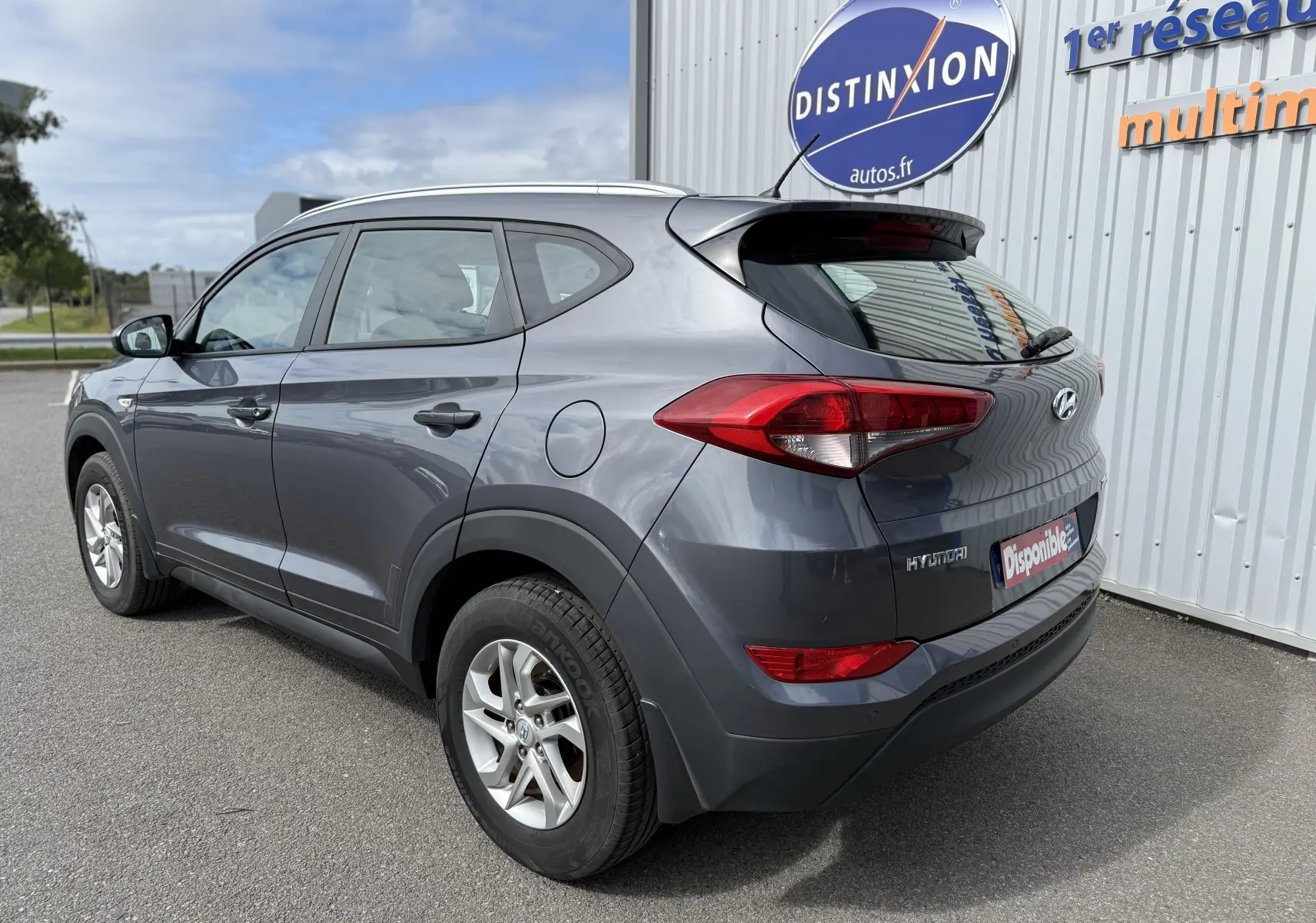 Vue 3/4 arrière droite d'un Hyundai Tucson gris 2015 stationné devant un bâtiment avec logo Distinxion.
