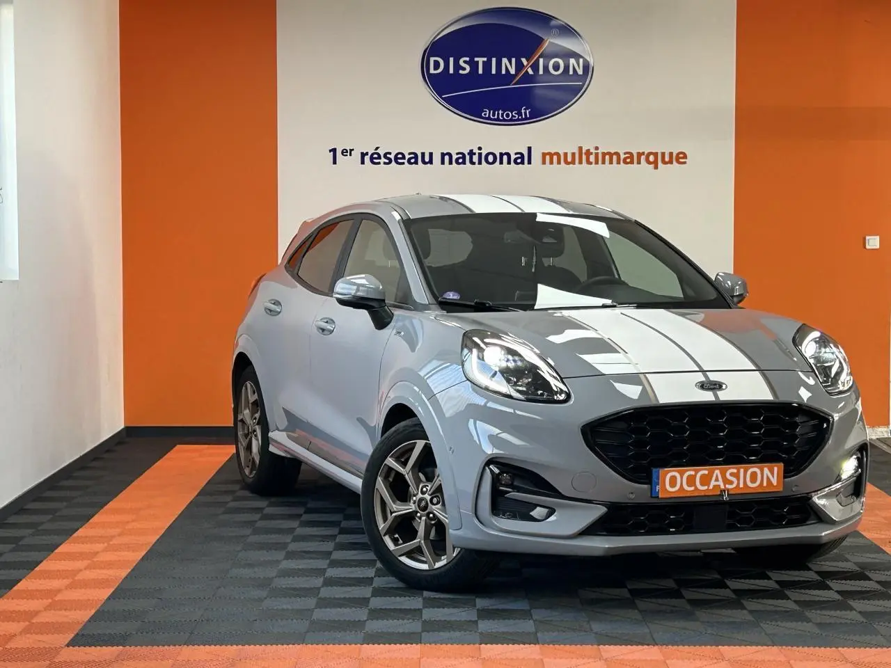 Vue 3/4 avant d'une Ford Puma gris clair avec bandes blanches, jantes alliage et calandre noire dans un showroom.