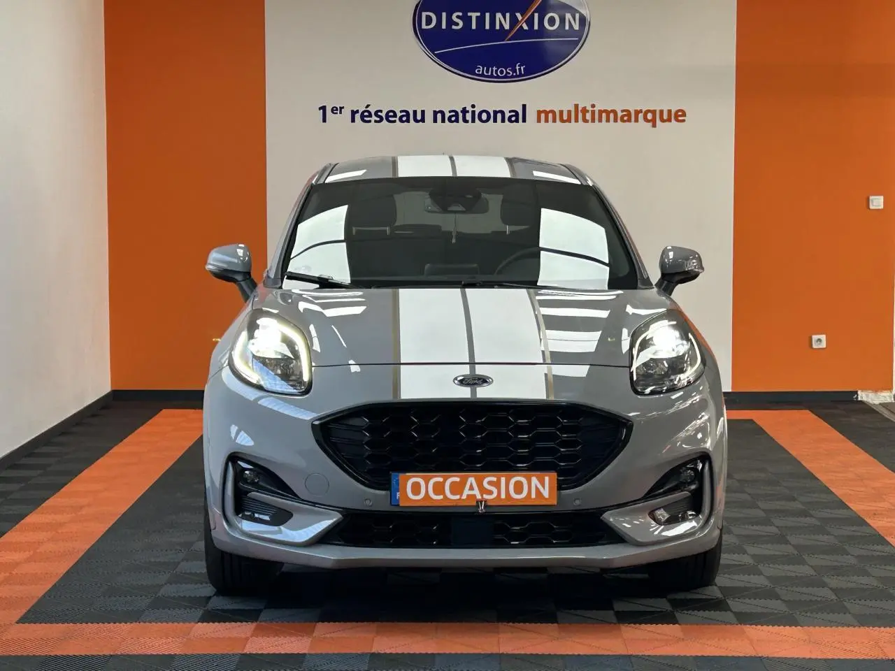Vue frontale d'un Ford Puma gris avec bandes blanches, phares allumés et plaque "OCCASION" orange.