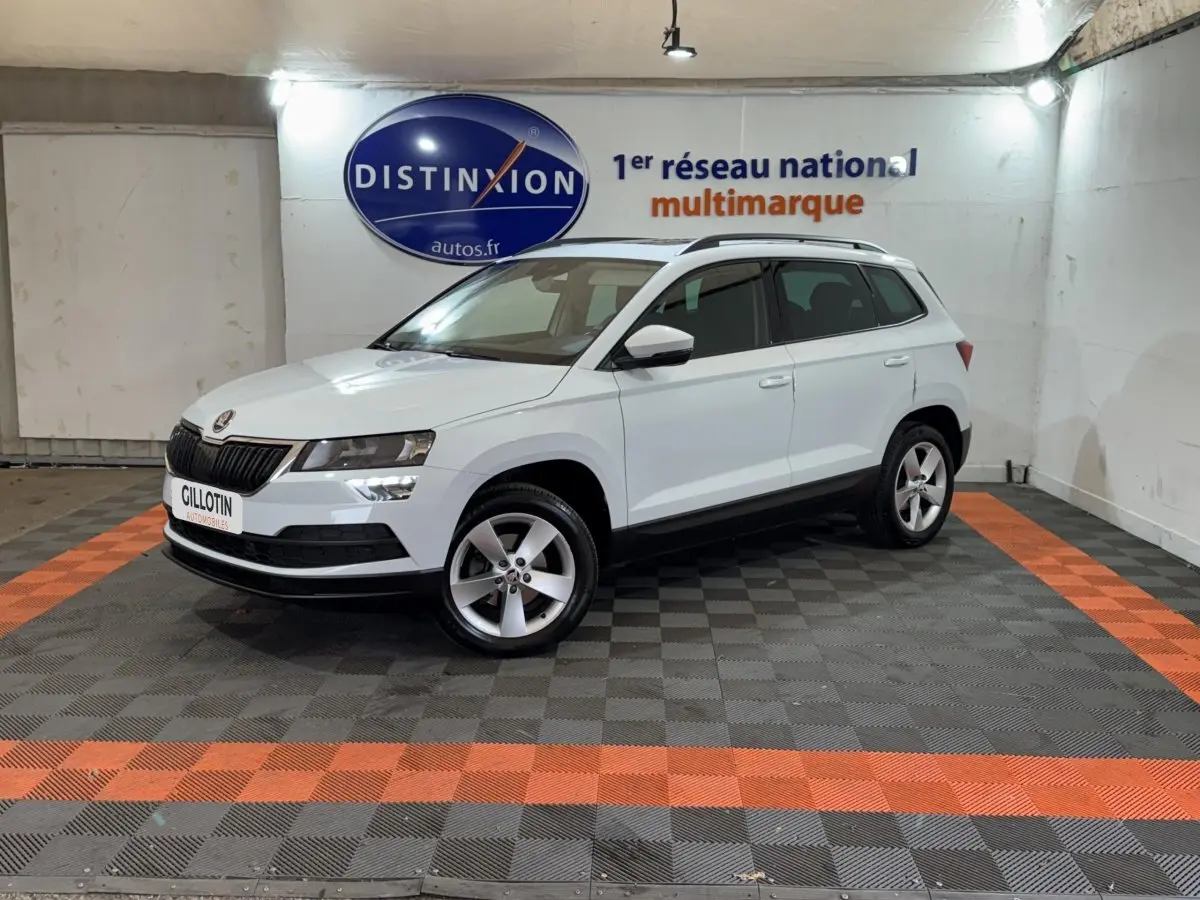 Vue 3/4 avant droite d'un Skoda Karoq blanc stationné en intérieur sur sol noir et orange avec logo Distinxion en arrière-plan.