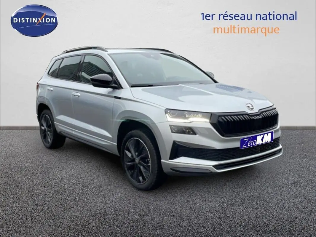 Vue 3/4 avant d'un Skoda Karoq argent 2025 avec barres de toit et jantes noires, sur fond neutre intérieur.