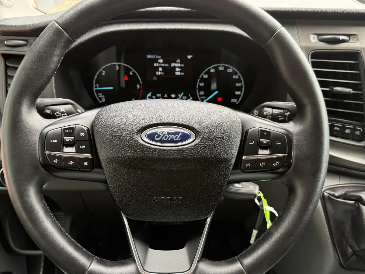 Vue rapprochée du volant en cuir noir du Ford Transit Fourgon 2024 avec commandes multifonctions et tableau de bord visible.