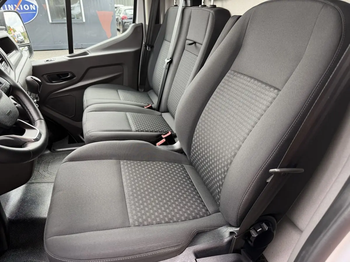 Vue intérieure côté conducteur du Ford Transit Fourgon 2024, montrant les sièges en tissu gris et le volant cuir.