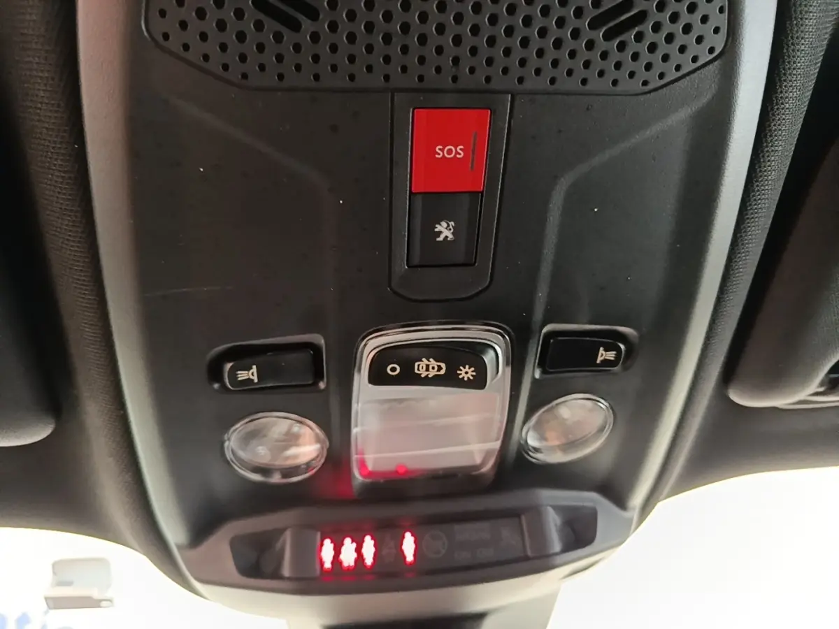 Vue en contre-plongée du panneau de commande de toit intérieur du Peugeot E-2008 électrique gris clair, avec bouton SOS rouge et éclairage.