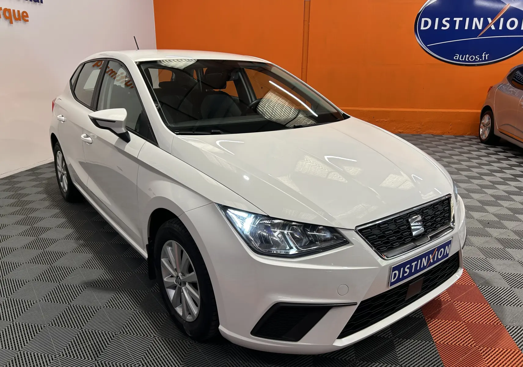 SEAT Ibiza blanche vue de 3/4 avant droit, avec phares LED allumés et calandre noire en intérieur showroom.