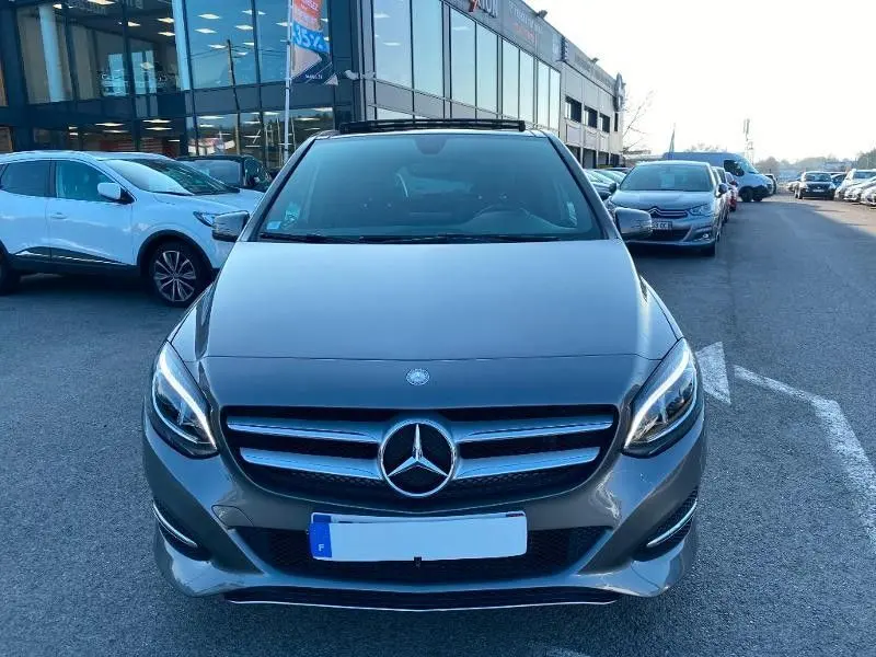 Vue frontale d'une Mercedes Classe B 180 d gris foncé métallisé de 2016, avec calandre distinctive et phares allumés.