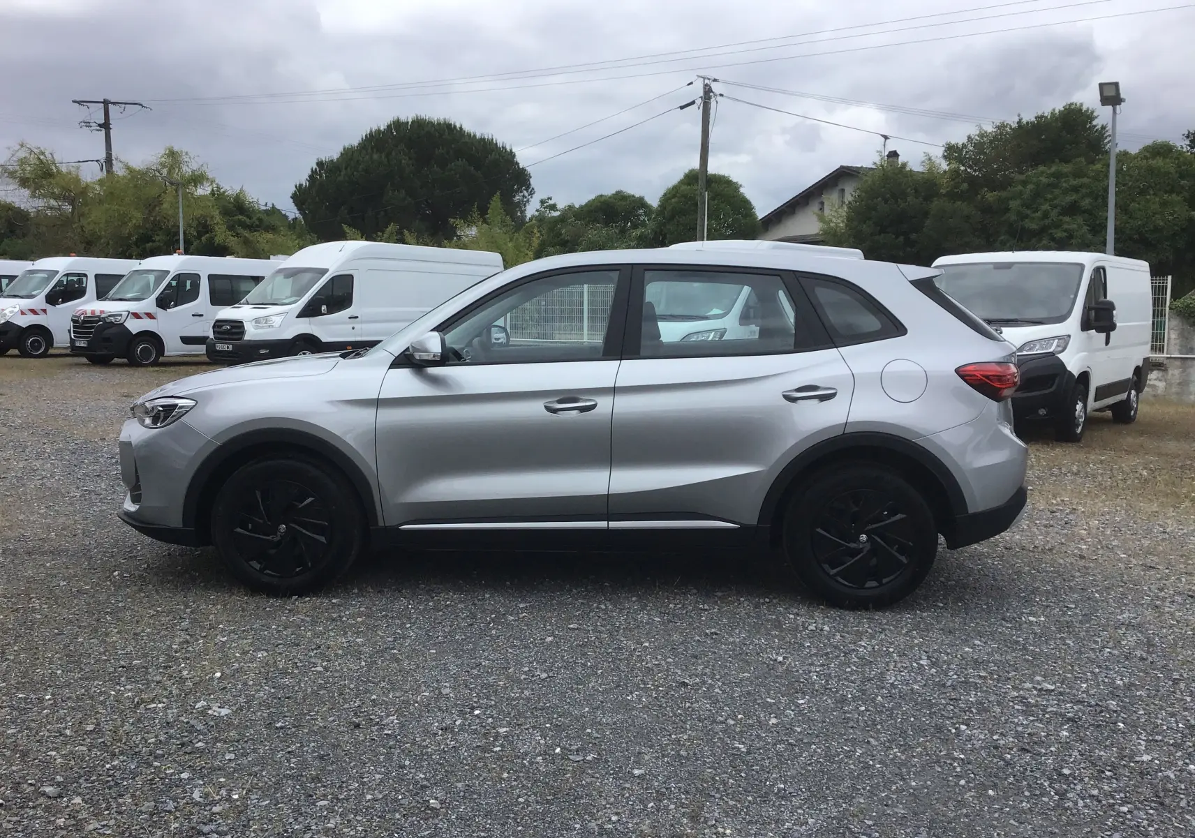 Vue de profil côté gauche du SUV MG ZS 1.5 L Hybrid+ 2026 en Cosmic Silver avec jantes noires sur un parking.