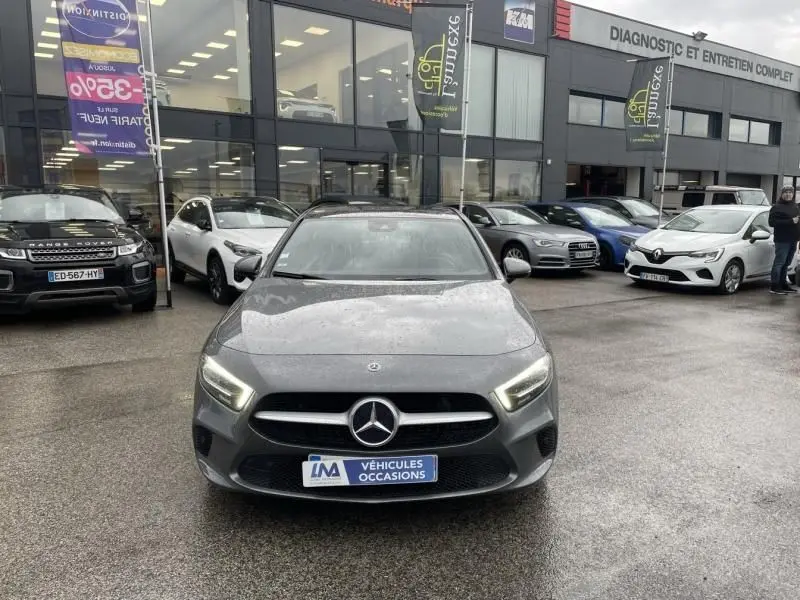 Vue de face d'une Mercedes Classe A 200 gris foncé métal, avec phares allumés, stationnée devant un garage.
