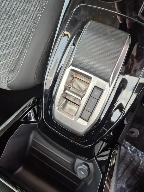 Gros plan sur la console centrale noire brillante de la Peugeot 208 Hybrid 110 Allure, avec sélecteur de boîte automatique et prise 12V.