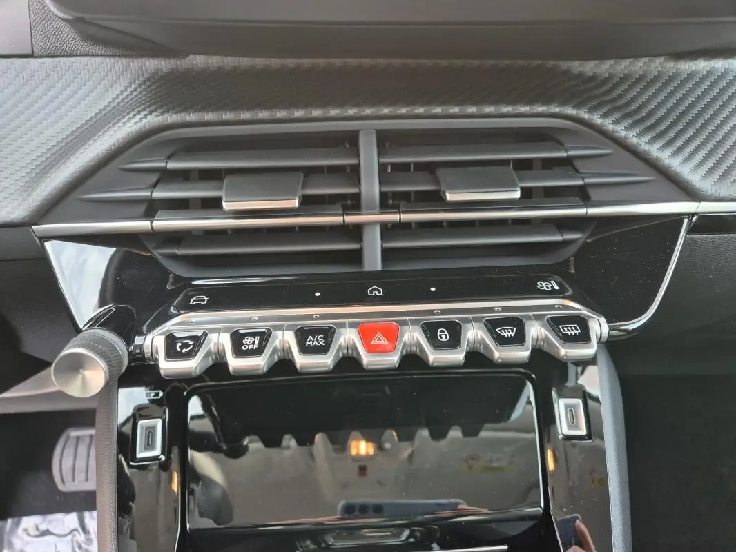 Gros plan sur la console centrale noire brillante de la Peugeot 208 Hybrid 110 Allure 2025, avec boutons de commande et aérateurs.