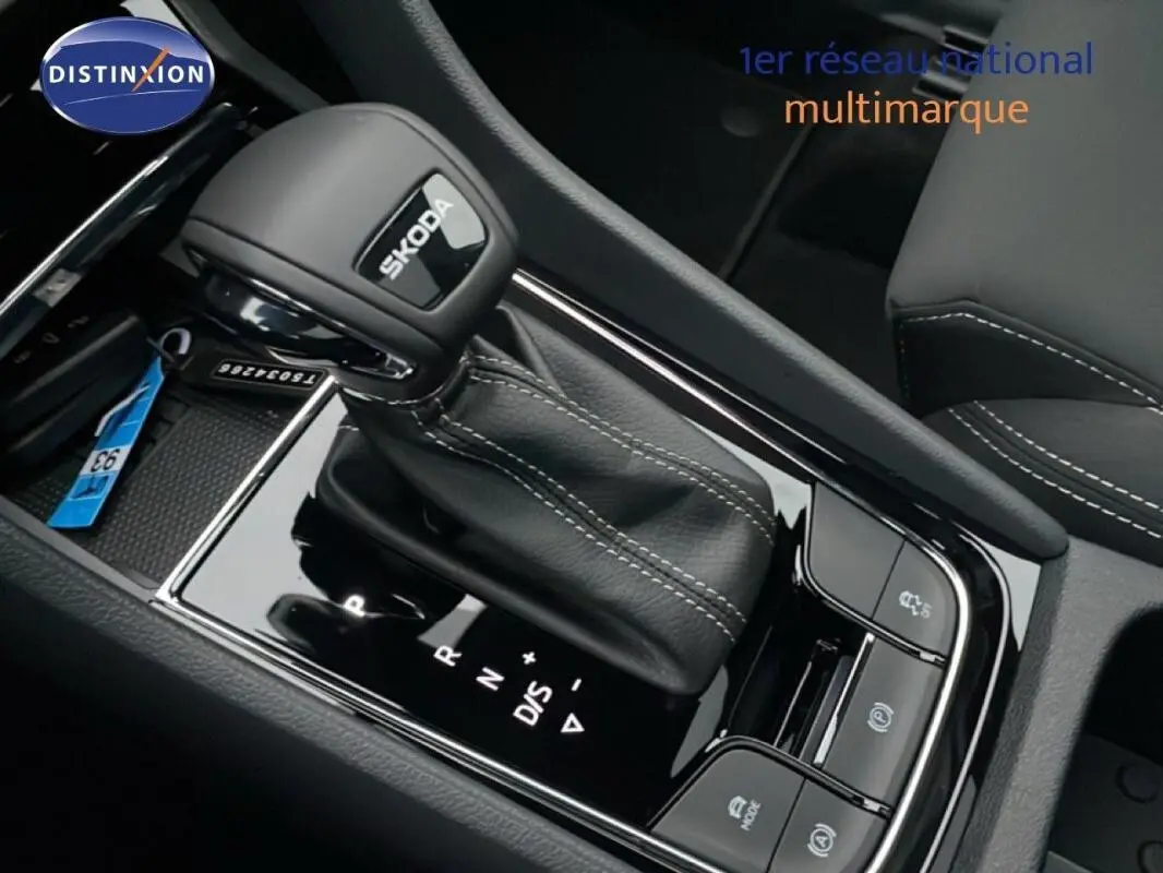 Gros plan sur le levier de vitesses automatique noir avec logo SKODA, entouré de commandes et coutures blanches.