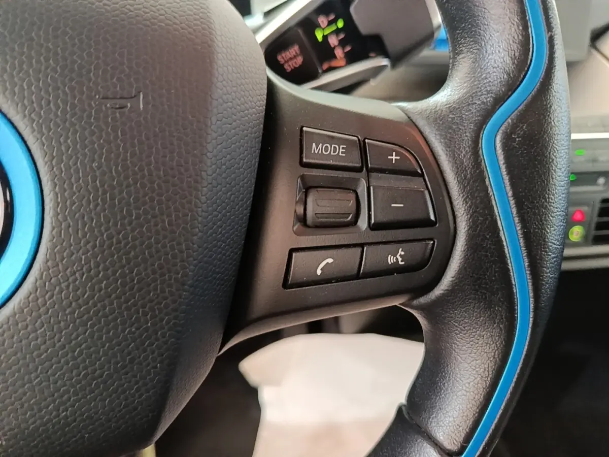 Gros plan sur le volant noir avec surpiqûres bleues de la BMW i3 120 Ah Atelier, montrant les commandes multifonctions.