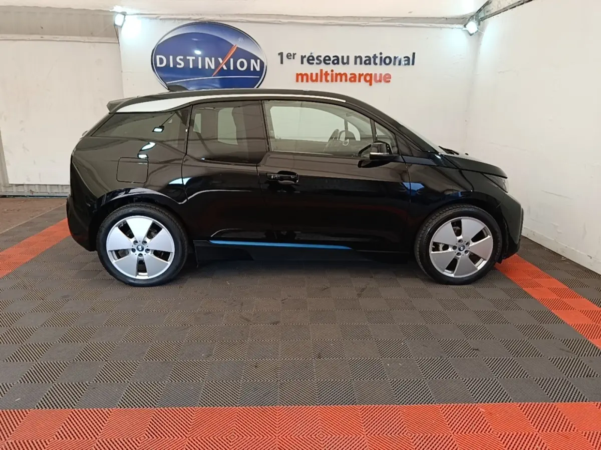 Profil côté gauche d'une BMW i3 120 Ah Atelier noire, compacte et électrique, stationnée en intérieur sur sol gris et rouge.