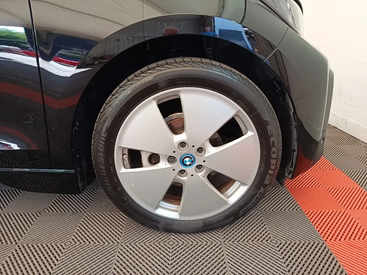 Gros plan sur la roue avant droite noire d’une BMW i3 120 Ah Atelier, avec jante argentée et logo BMW central.