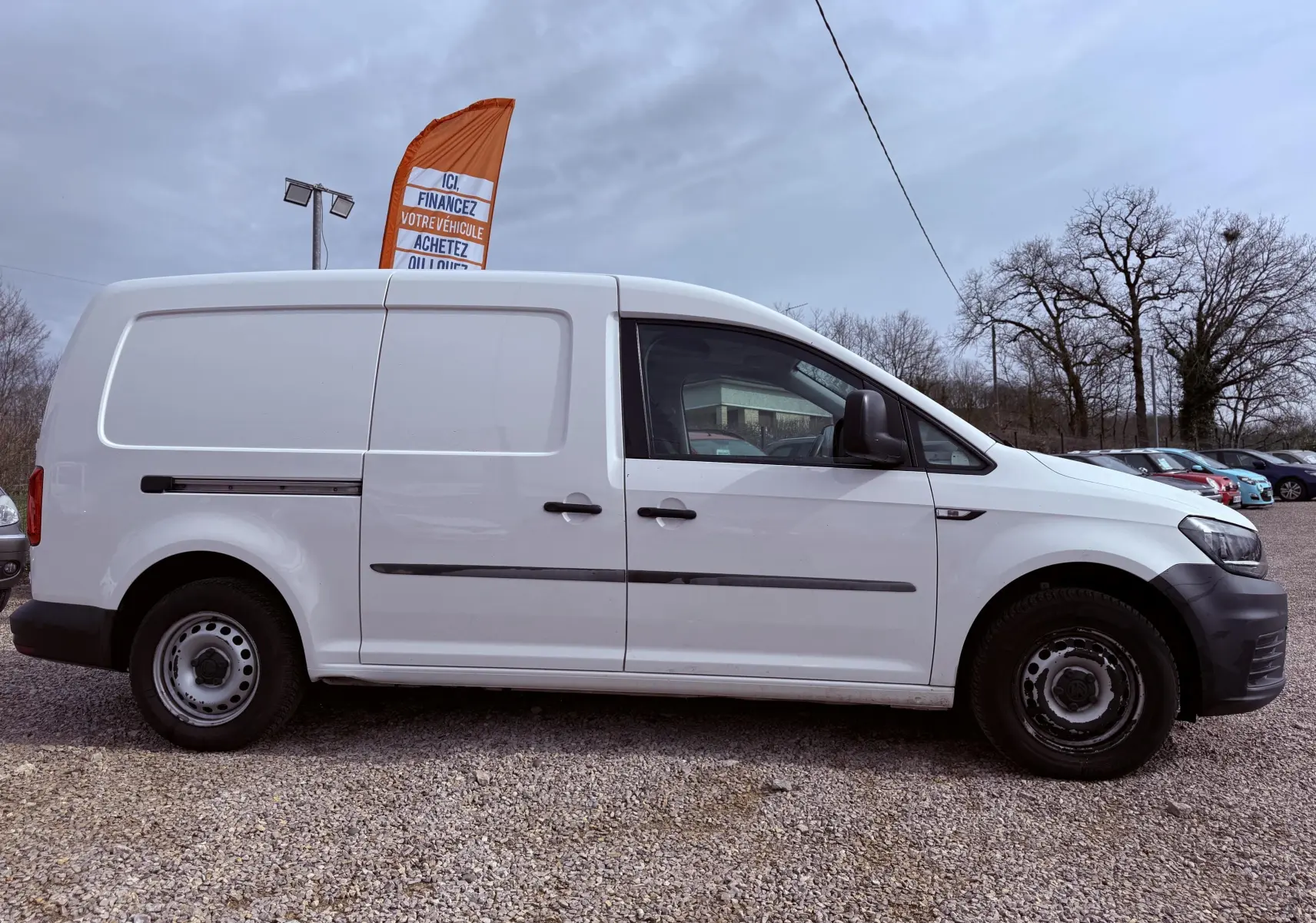 Vue latérale gauche d’un utilitaire Volkswagen Caddy Maxi Long blanc avec pare-chocs noirs sur un parking extérieur.